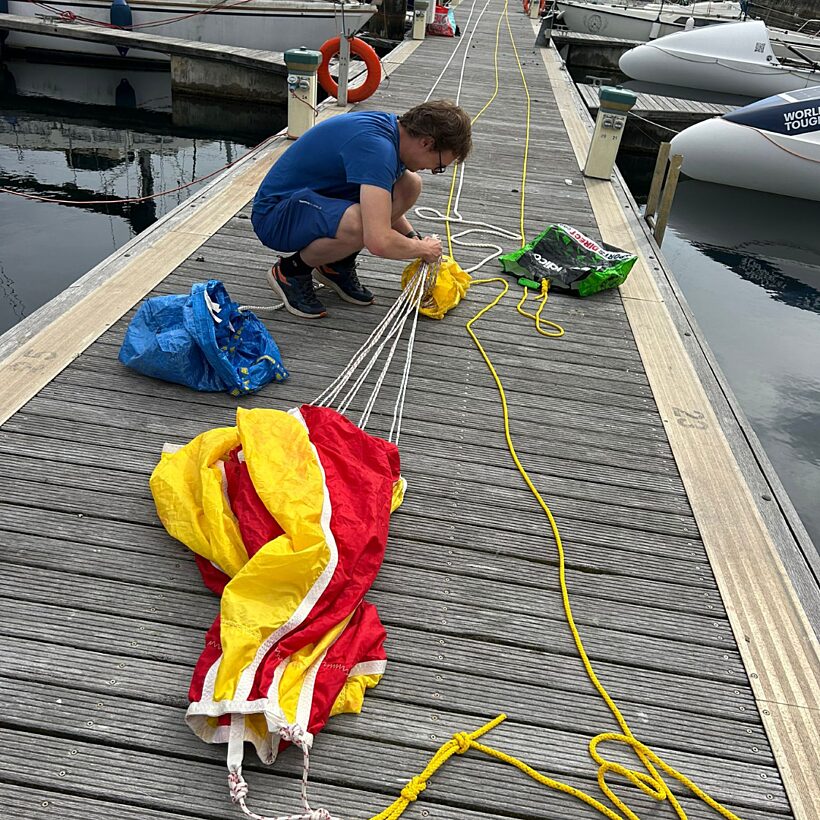 Man in blue top crouched on wooden deck folding a yellow and red parachute