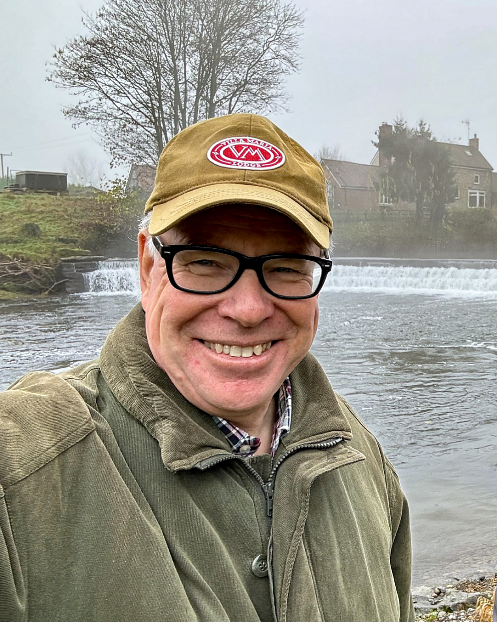 White man in green jacket and yellow hat stood in front of river and weir