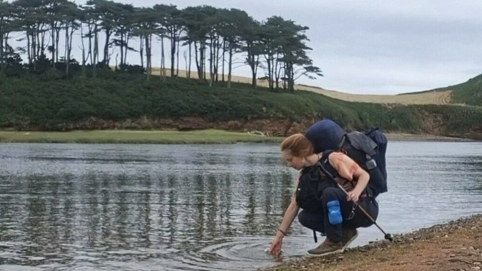 A woman with a backpack bends down to dip her hand in a low river