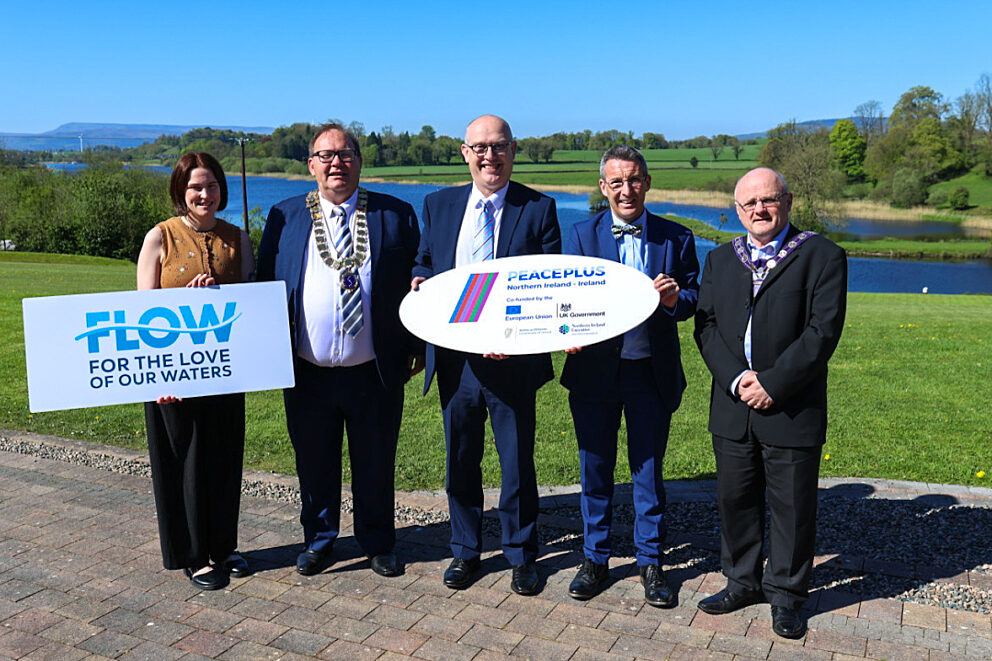 Group of five people, four men and one woman stood in suits in front of a river holding signs reading FLOW.