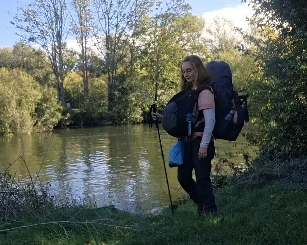 a woman with a backpack and walking pole sands next to a river looking at the water
