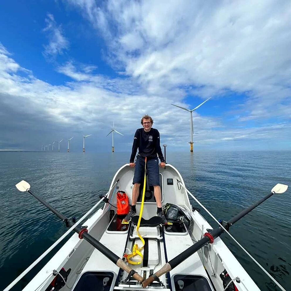 A man stands on a white boat with oars in a calm ocean with blue skies and a windfarm visible behind