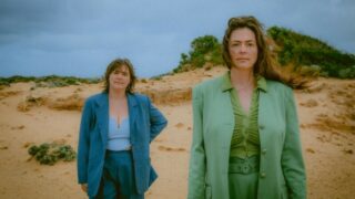 A photograph of two women wearing blue and green suits standing on a beach, looking at the camera