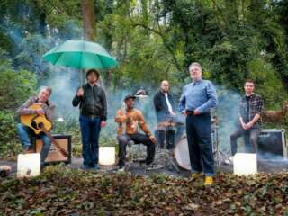 Six men, the band China Crisis, stand and sit with their instruments, surrounded by trees