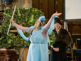 A musician in a blue dress stands in front of a microphone, arms outstretched, holding a flute