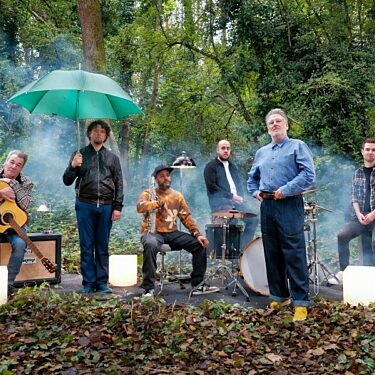 Six men, the band China Crisis, stand and sit with their instruments, surrounded by trees