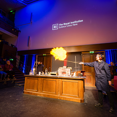 Photograph of the stage with a desk with one person demonstrating a fire experiment