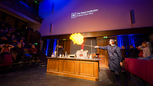 Photograph of the stage with a desk with one person demonstrating a fire experiment