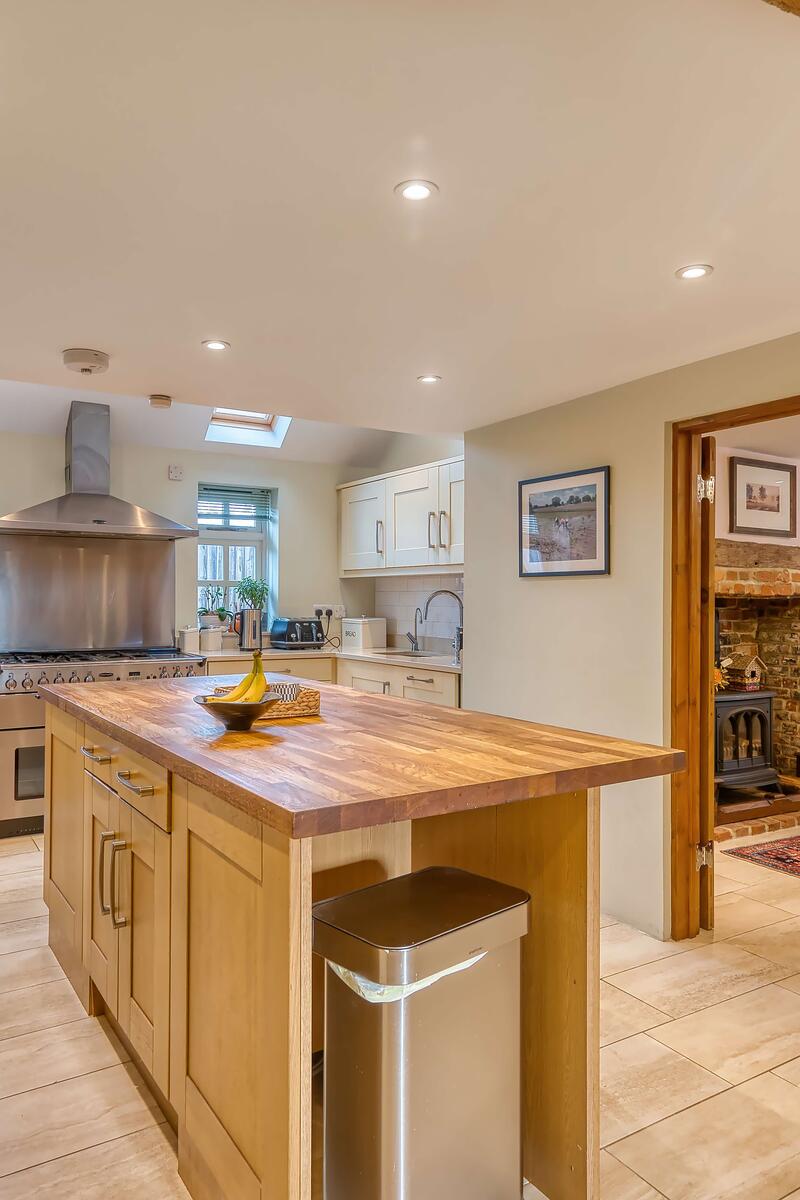 Bright farmhouse kitchen with central wooden island, cream cabinetry, exposed ceiling beam and open doorway leading to a dining room with wood burner.