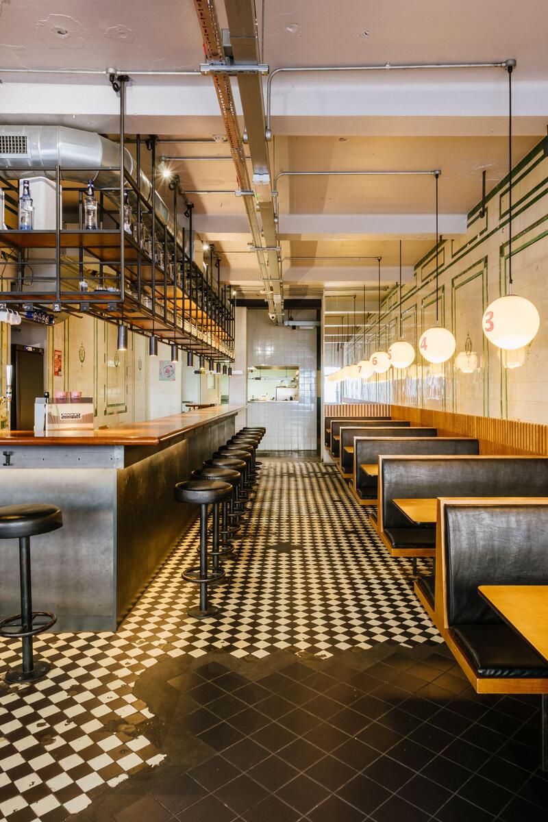 Industrial restaurant interior with checkerboard floor, timber booth seating, tiled walls and long metal bar in East London shoot location
