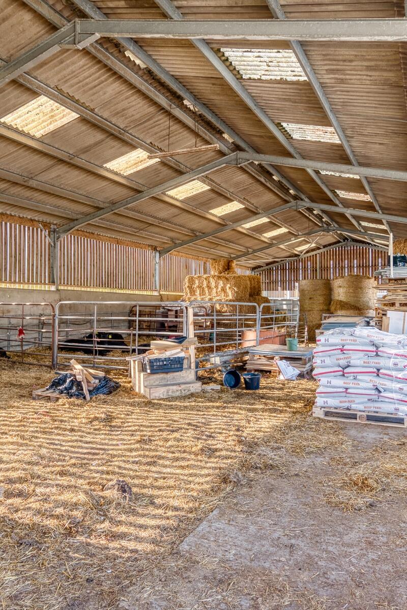 Large open agricultural barn with exposed steel frame, corrugated roof panels, hay bales and livestock pens, ideal rural farm filming location.