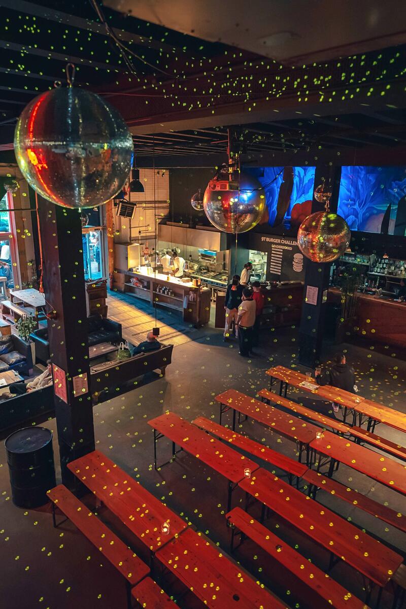 Large industrial bar and event space with wooden benches, long bar counter, disco balls and atmospheric lighting.