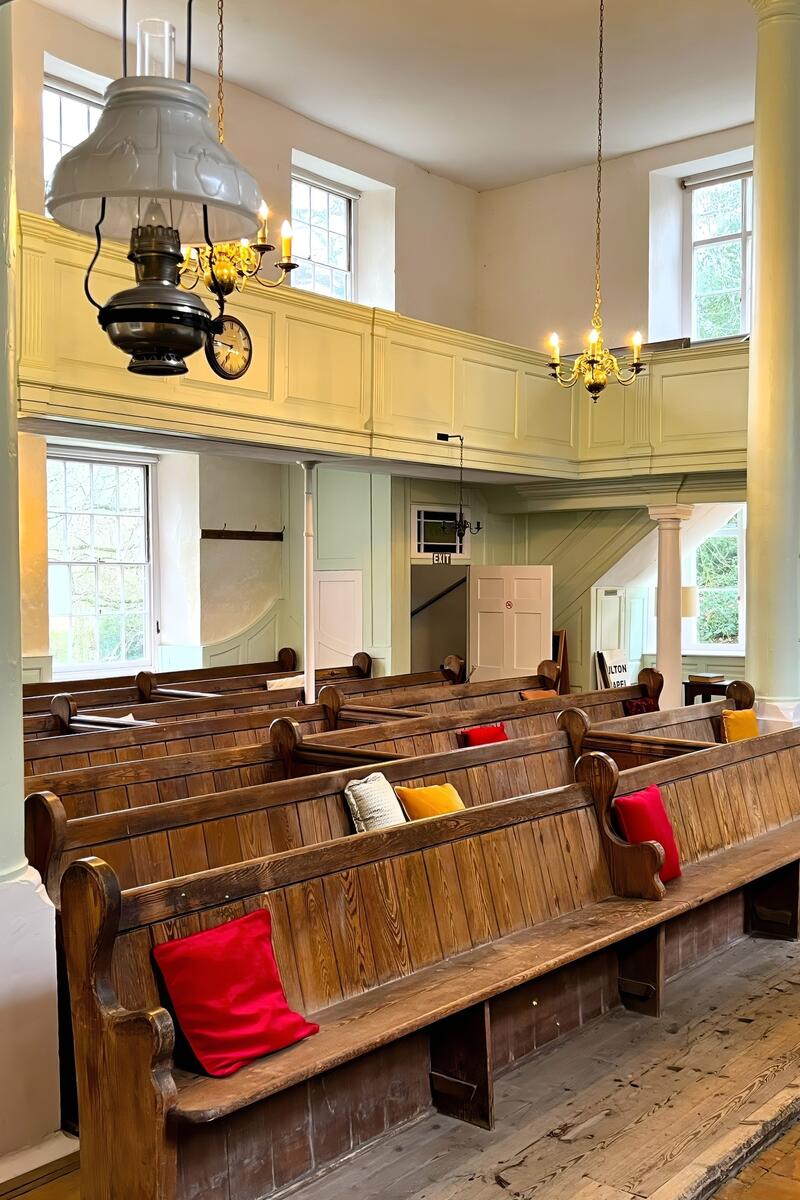 Interior of a historic country church with wooden pews, pale green gallery balcony, tall windows and traditional hanging lamps.