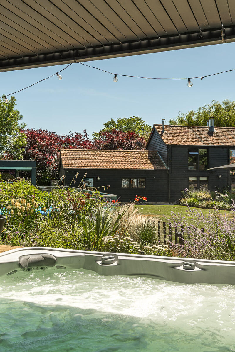 Covered outdoor hot tub overlooking landscaped gardens and a luxury black barn conversion with countryside views.