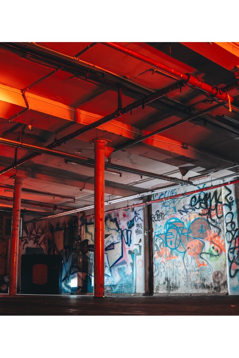 Large industrial warehouse space in a former Manchester textile mill with graffiti-covered walls, exposed steel columns, pipework and red lighting.