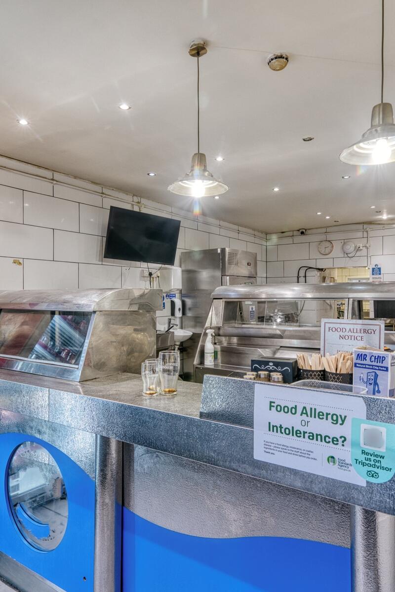 Traditional London fish and chip takeaway with stainless steel serving counters, glass food displays, drinks fridge and white tiled walls.