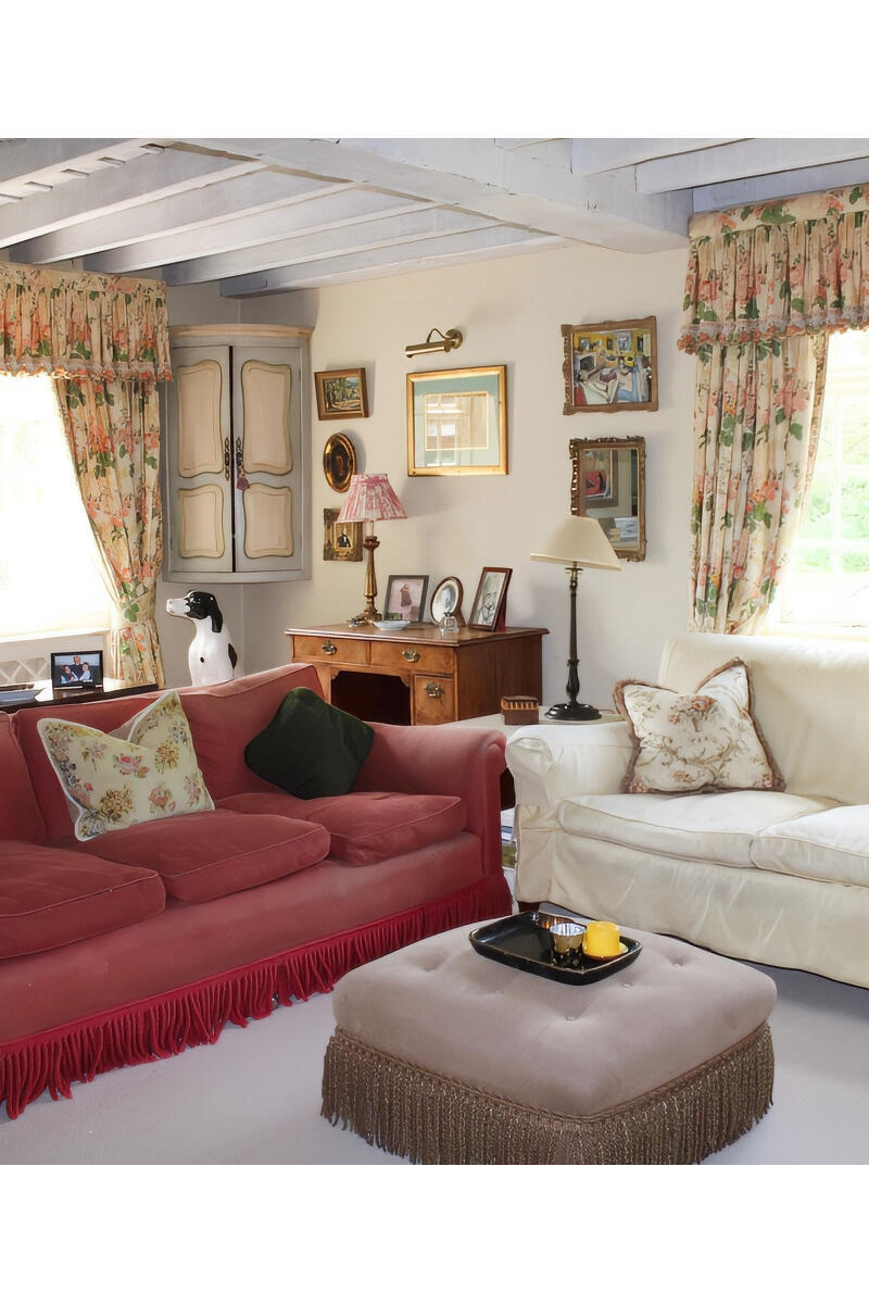 Traditional country sitting room with floral curtains, exposed beams, a red fringe-trim sofa, a white sofa, and a central ottoman, styled with vintage furniture and framed wall art.