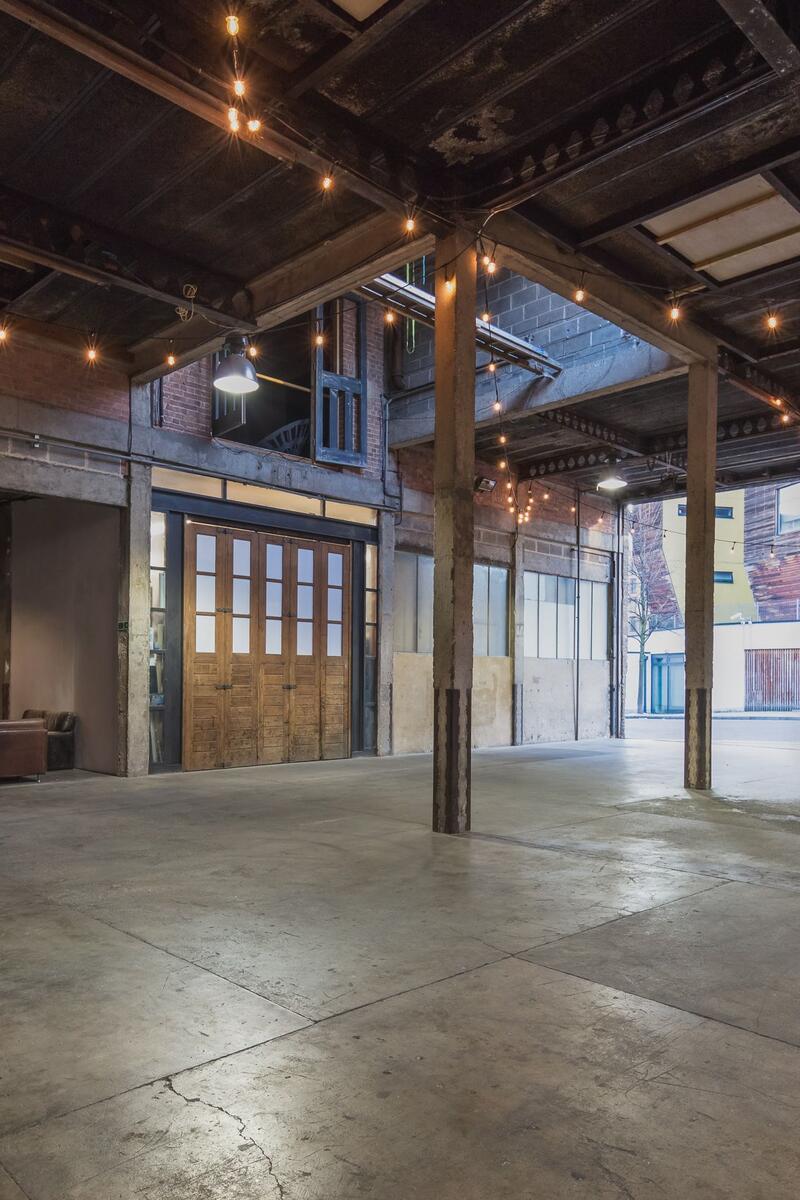 A large, industrial-style event space or warehouse venue, featuring exposed brick walls, concrete floors, high ceilings with visible structural beams, and string lights illuminating the space.
