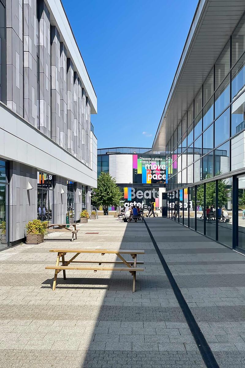 Modern university campus courtyard in Hertfordshire with contemporary glass buildings, outdoor seating, wide pedestrian walkway and clear blue sky.