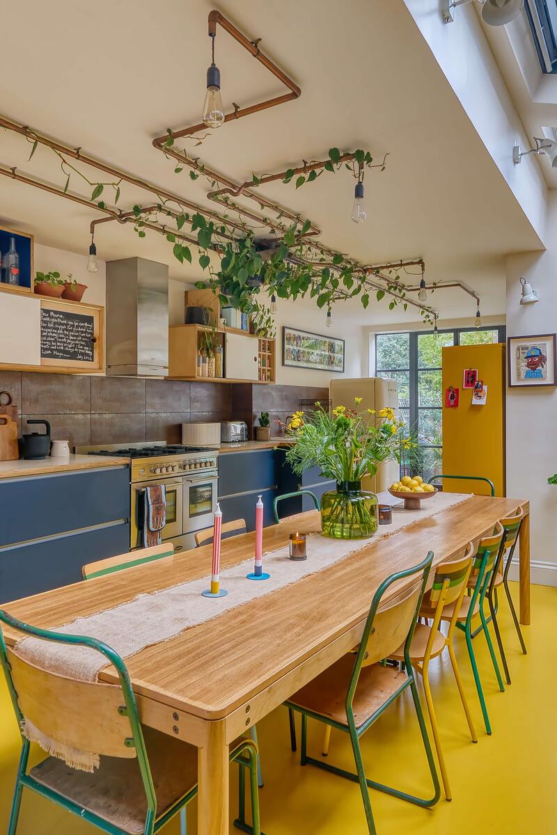 Colourful industrial-style kitchen with bold yellow floor, exposed pipework, skylights, and plants creating a vibrant shoot location.