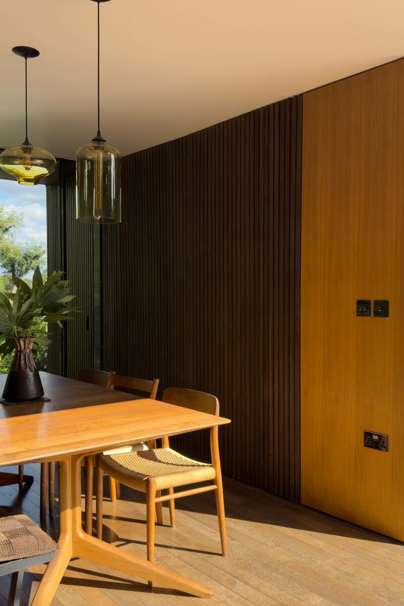 Modern dining area with a wooden table, mixed seating of chairs and bench, and pendant lights hanging above. Large floor-to-ceiling windows open onto greenery, filling the space with natural light.