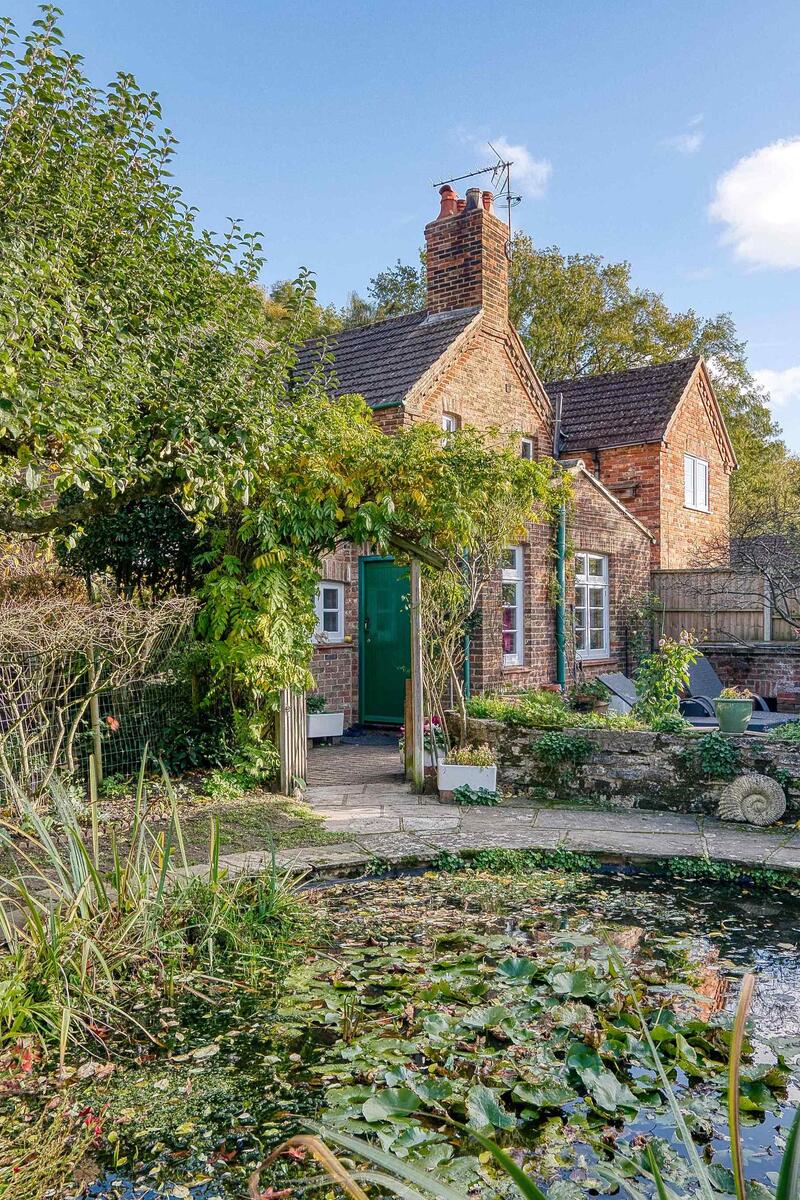 Red-brick period cottage in Surrey set behind a mature garden with pond, trees and winding path, offering a natural outdoor setting ideal for filming and photography.