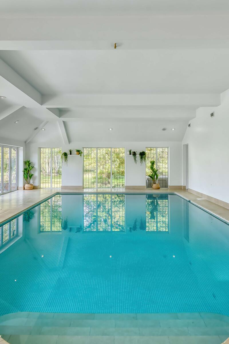 Bright indoor swimming pool with tiled flooring, full-height windows, vaulted white ceilings and garden views throughout