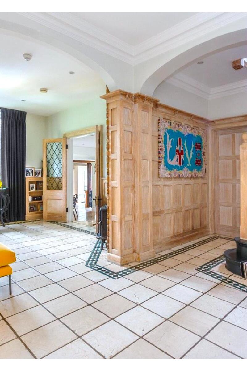 A large Tudor-style entrance hall with pale wood panelling, tiled flooring with green inlay detailing, a striped staircase, and an arched opening leading to adjoining rooms.