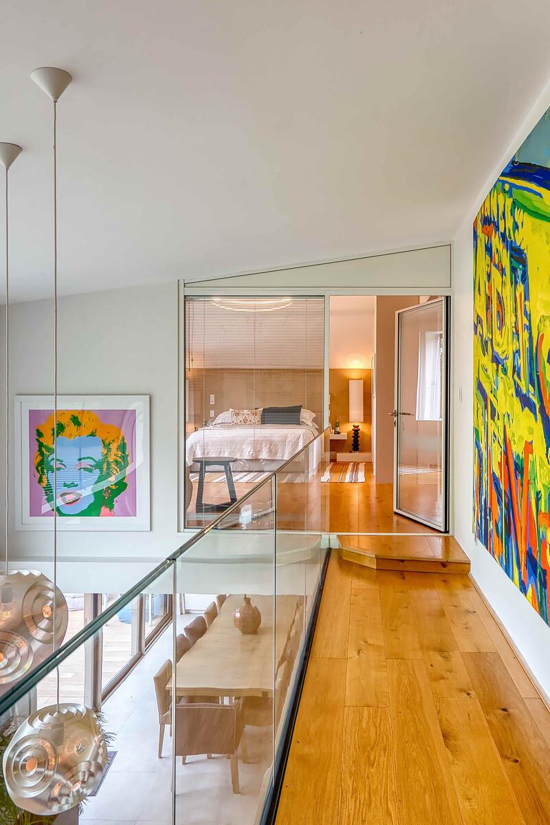 Bright first-floor landing with glass balustrade, large modern artworks, pendant lighting and a view into a contemporary bedroom.