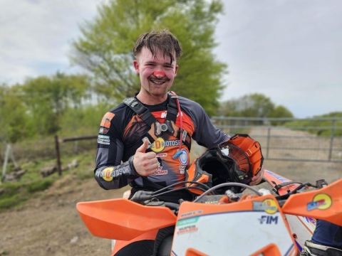 An offroad motorcyclist rests and celebrates with a thumbs up at the end of a course. The rider looks hot and sweaty having removed his helmet against a rural backdrop.
