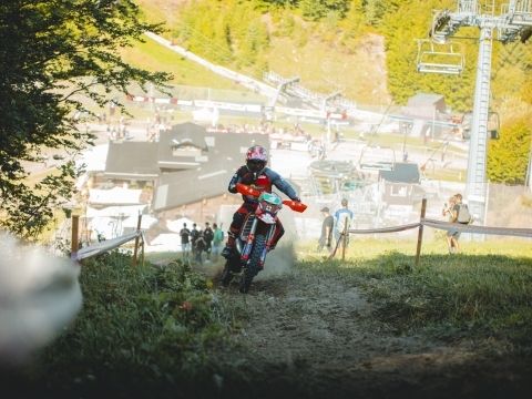 Enduro rider accelerating uphill on an orange KTM enduro motorcycle heading towards a shaded area of tree.