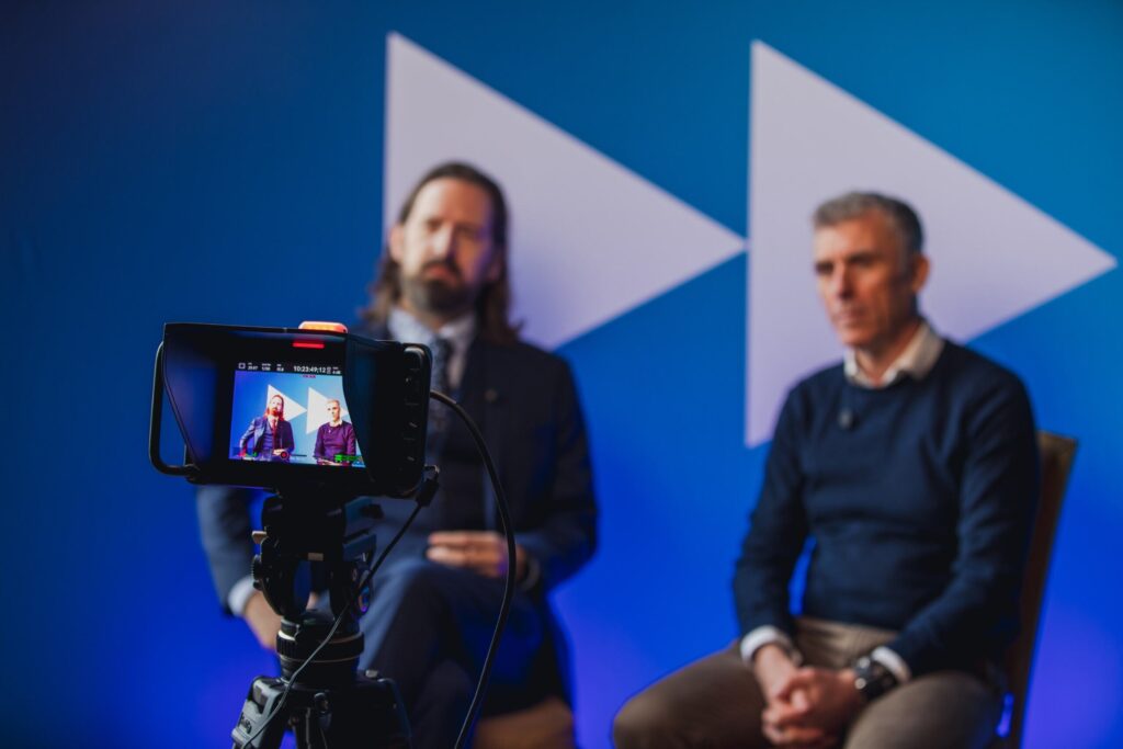 Two people in camera, sitting against blue backdrop with big white arrows