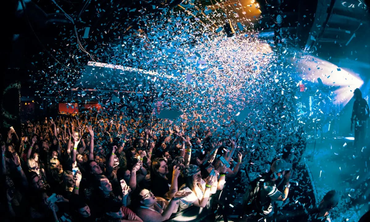 Crowd at a concert with arms raised as blue confetti falls under stage lighting.