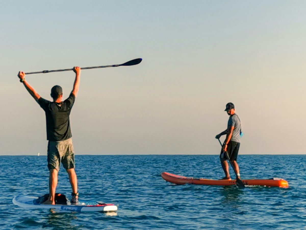 Two lads stand-up paddleboarding on a calm sea at sunset, one with his paddle raised in the air