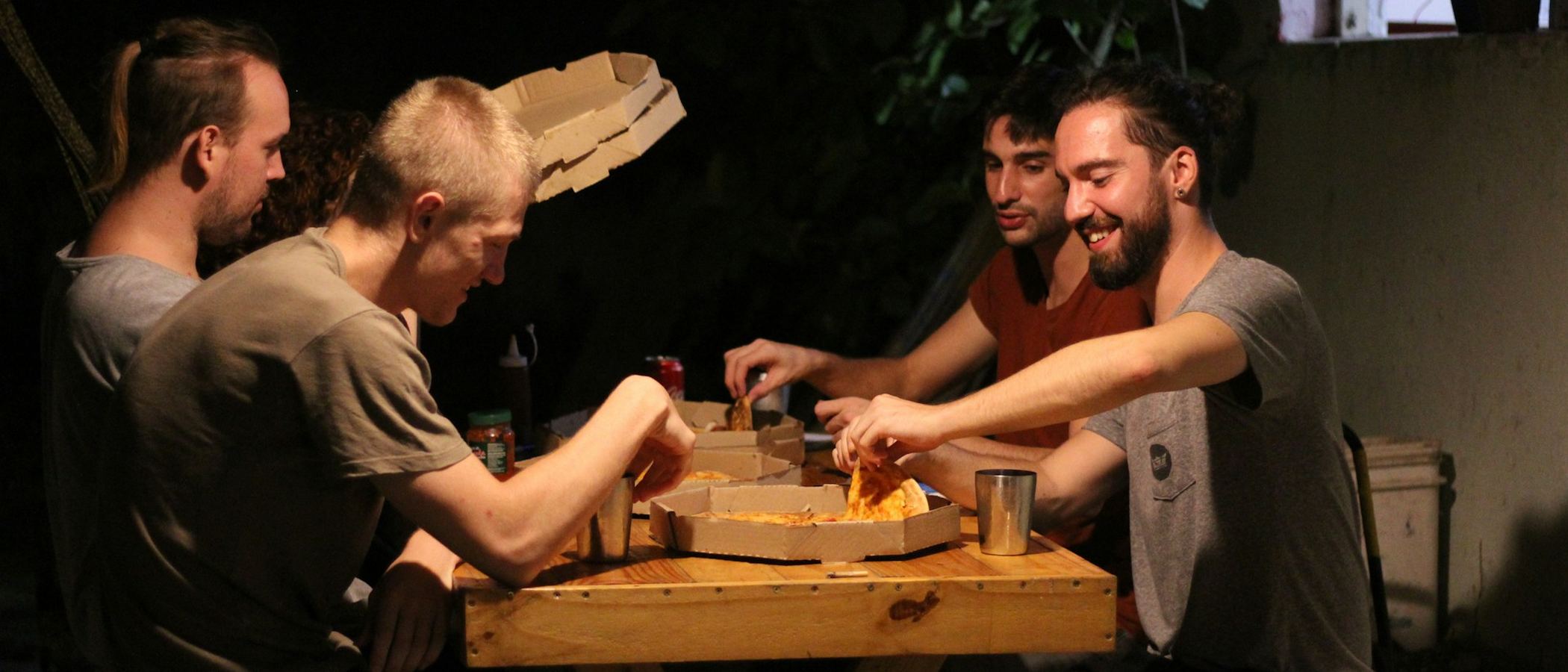 Group of lads laughing and sharing pizza around an outdoor table at night