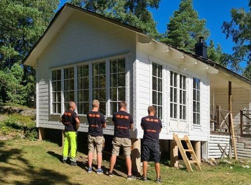 Fyra hantverkare i L&A-tröjor står framför vitmålad sommarstuga med många fönster, solig dag
