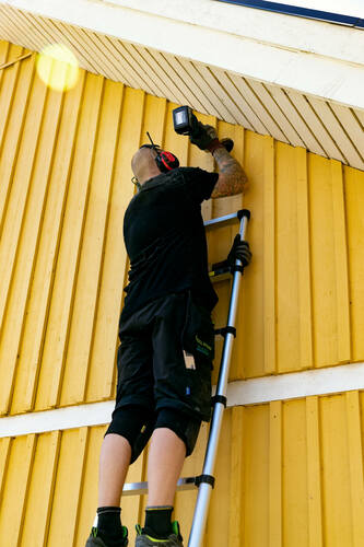 Tekniker installerar ventilationsgaller på vindskiva från stege vid gul träfasad
