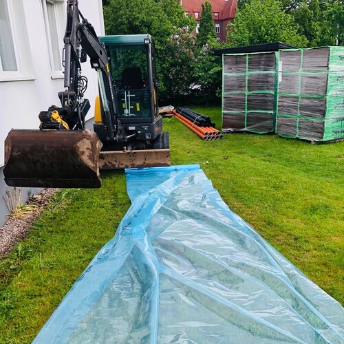 Grävmaskin i arbete vid villa inför dränering, med skyddsplast och byggmaterial redo