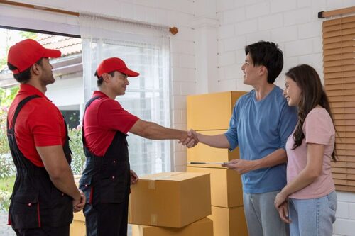 Två flyttare i röda uniformer som skakar hand med kunder i ett hem fyllt av kartonger.