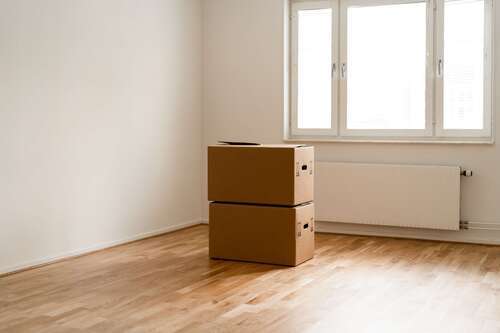 Två flyttkartonger staplade i ett tomt rum med trägolv och stort fönster.