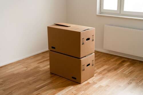 Två staplade flyttkartonger i ett tomt rum med trägolv och fönster i bakgrunden.