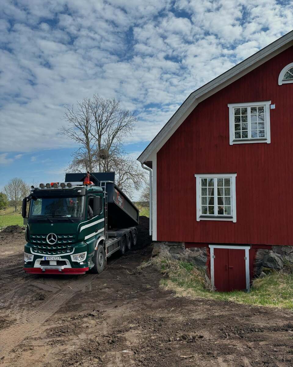 Tung lastbil tippad med jord intill röd lada – leverans av fyllnadsmassor för tomtplanering i Uppsala