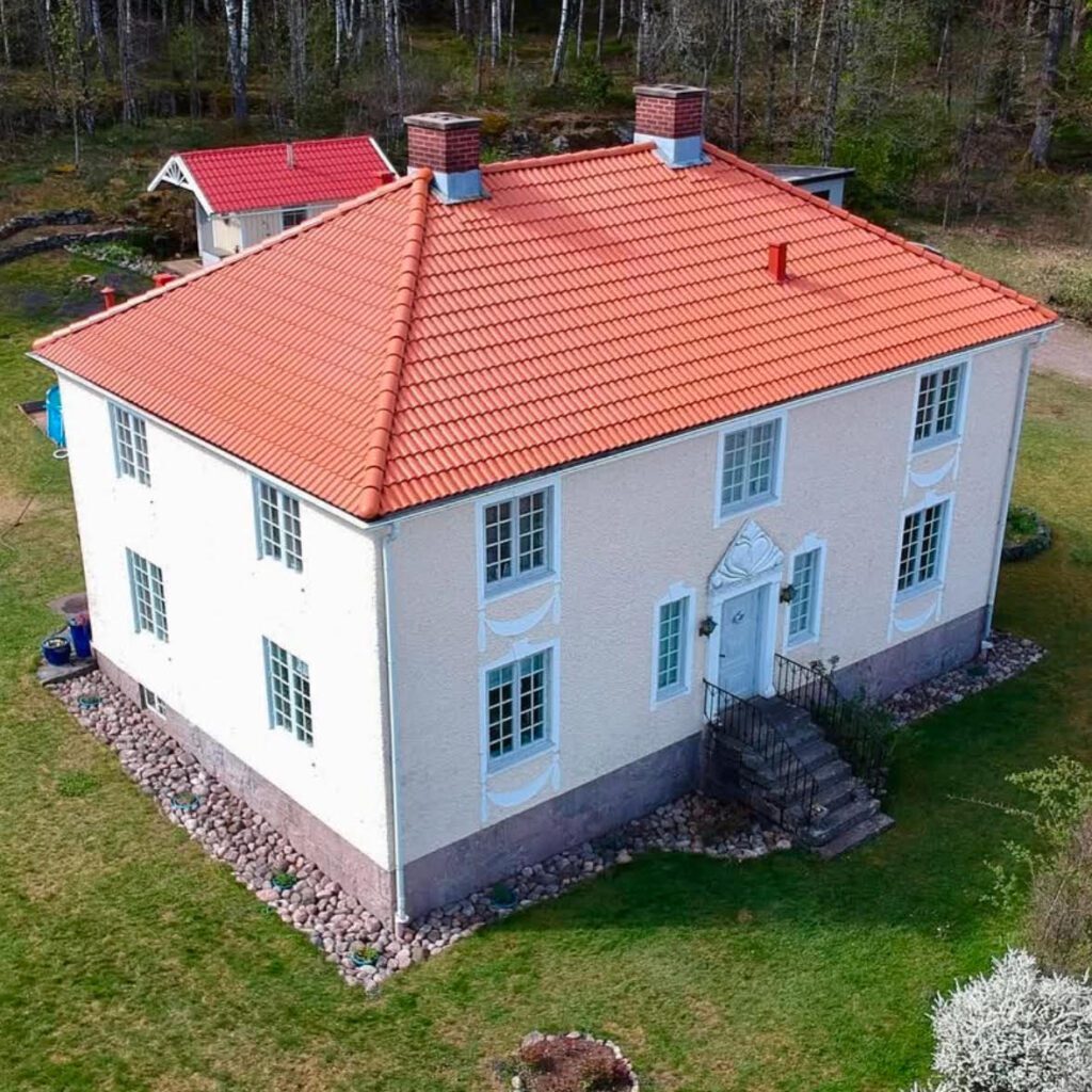 Ljus putsad tvåplansvilla med nytt rött tegeltak och valmat tak, fotograferad uppifrån i grönskande omgivning.