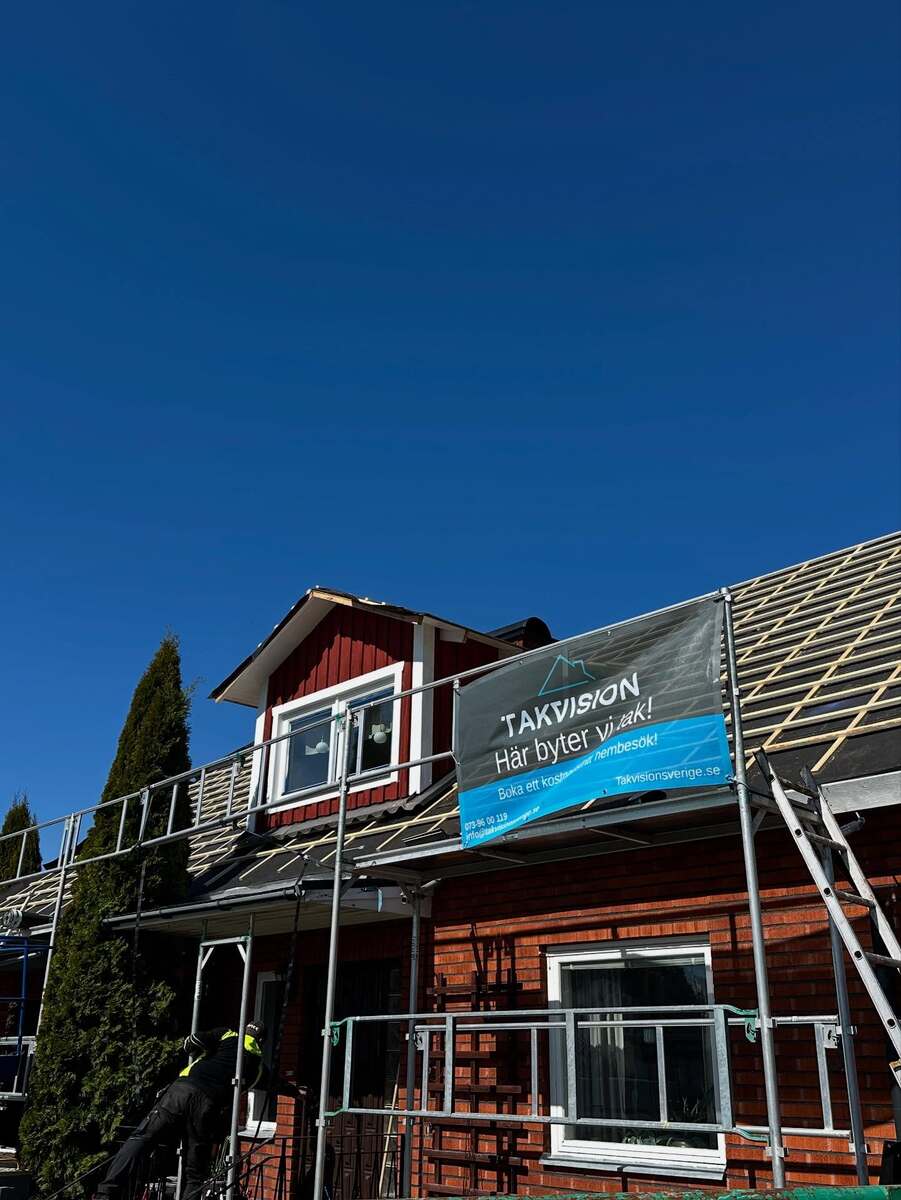 Pågående takarbete på villa med rött tegel och frontespis, byggställningar och Takvision-banderoll tydligt synliga under klarblå himmel.