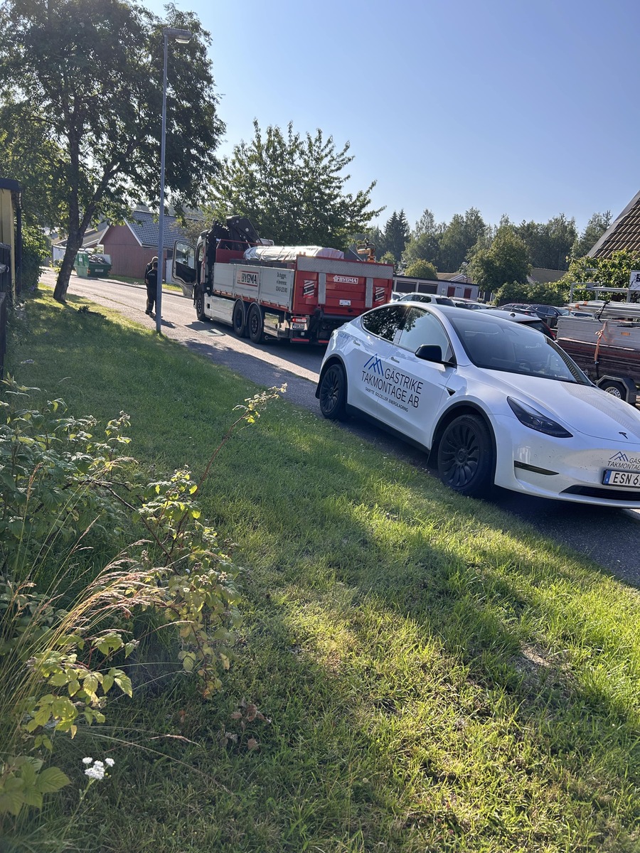 Vit bil med företagslogotyp parkerad vid villaområde, lastbil med material i bakgrunden