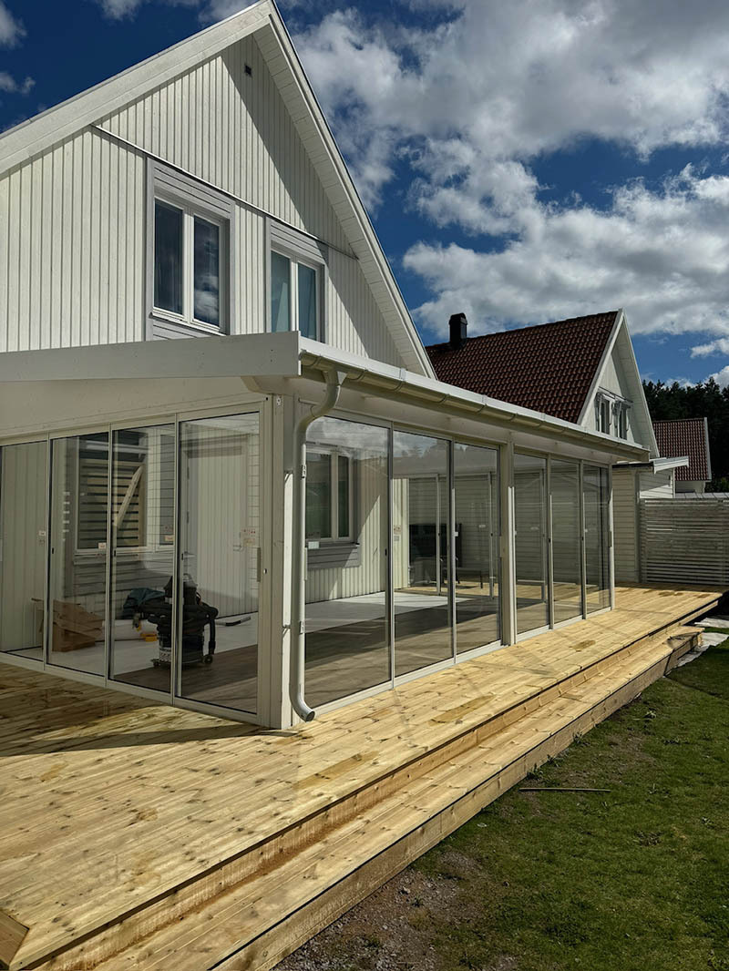 Vitt hus med inglasat uterum och nybyggd altan i trä under blå himmel.