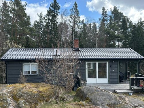 Ett grått fritidshus med svart tak och skorsten i skogsmiljö.