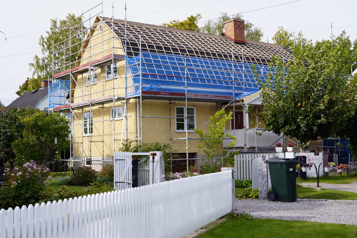 Villa med byggställning runt taket under takrenovering, vit staket och trädgård i förgrunden.