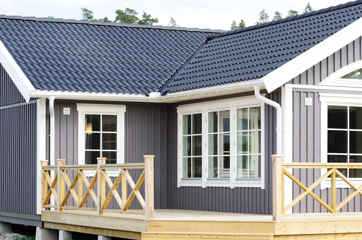 Modernt hus med blå fasad, vita fönster, nytt tak i mörk takpanna och altan med trästaket.