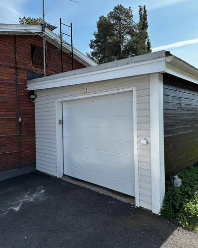 Fristående garage med vit port och träpanel, placerat intill ett rött tegelhus.